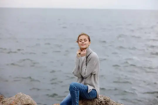 Молодая женщина с закрытыми глазами сидит на камне у моря
