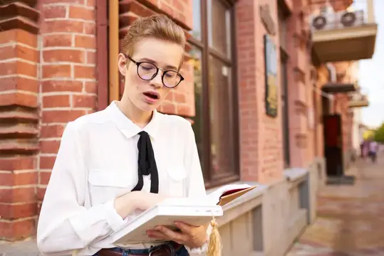 Рыжеволосая девушка в очках с книгой у кирпичного дома