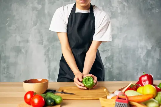 Повар в чёрном фартуке режет зелёный перец на кухне