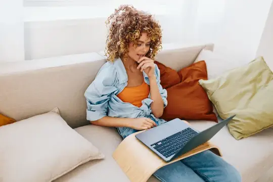 Кудрявая девушка с ноутбуком сидит на диване с подушками