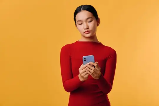 Женщина с длинными черными волосами в красном топе использует смартфон для обмена сообщениями или просмотра веб-страниц на однотонном оранжевом фоне