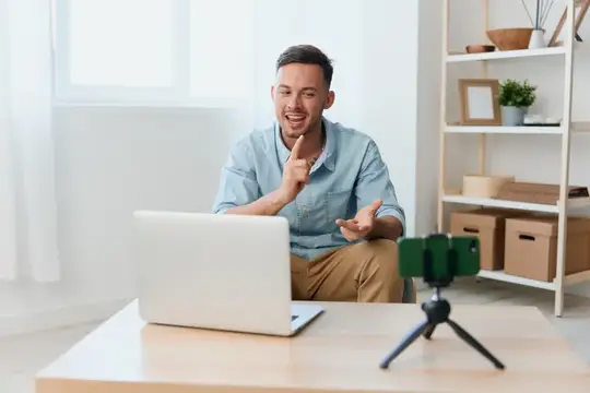 Улыбающийся мужчина записывает видео на смартфон перед ноутбуком