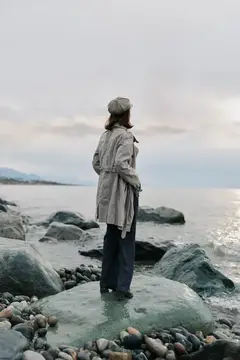 Женщина в плаще и берете стоит на камнях у моря