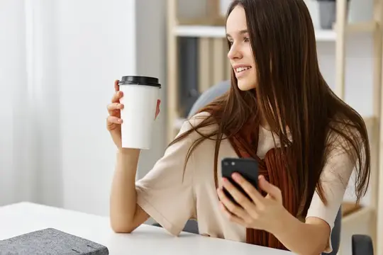 Улыбающаяся девушка с кофе смотрит в смартфон за столом