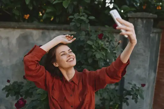 Яркая, расслабленная женщина в повседневной одежде позирует на улице, улыбаясь в телефон, пока рядом цветут цветы