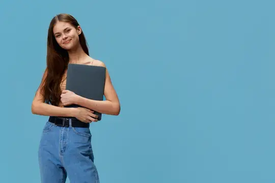 Улыбающаяся молодая женщина с длинными каштановыми волосами, держащая ноутбук в повседневной одежде, стоит уверенно с расслабленной позой на фоне однотонного голубого фона, излучая непринужденность…