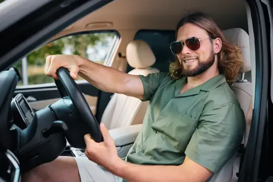 Улыбающийся длинноволосый мужчина в солнцезащитных очках за рулём автомобиля
