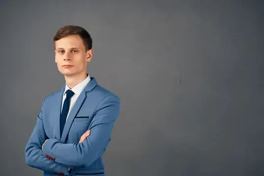 Молодой человек в синем костюме со скрещёнными руками на сером фоне