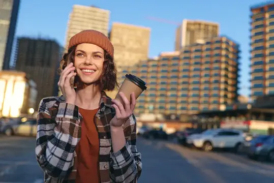 Улыбающаяся девушка в шапке говорит по телефону с кофе на фоне города