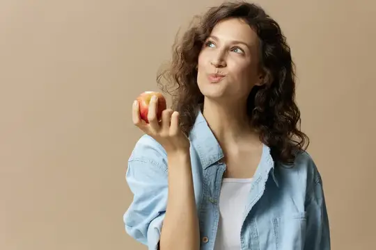 Кудрявая женщина с яблоком мечтательно смотрит вверх на бежевом фоне