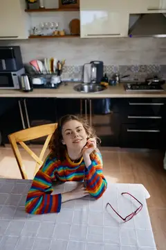 Молодая женщина в разноцветном свитере сидит за столом на кухне