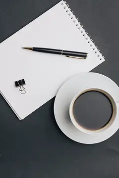 На столе лежит блокнот, стоит чашка кофе и ручка — офисная работа, бизнес