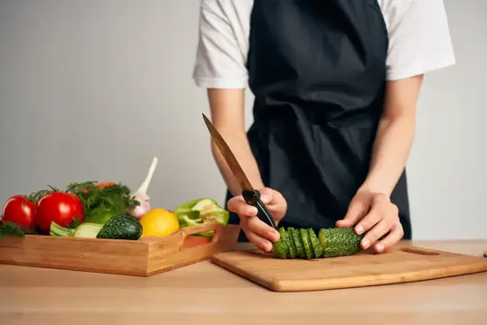 Повар в чёрном фартуке нарезает огурец на доске рядом с овощами