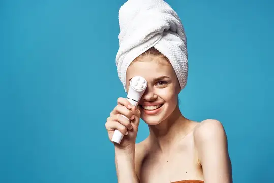 Улыбающаяся светловолосая девушка с полотенцем на голове и массажером для лица