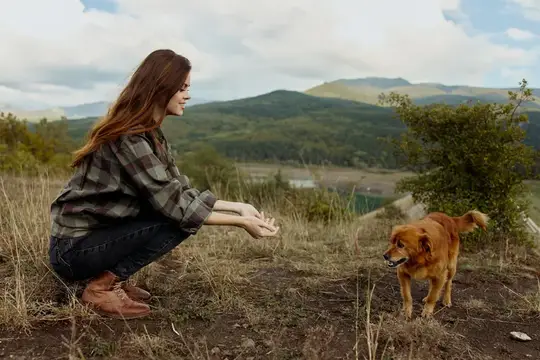 Девушка присела и зовёт рыжую собаку на фоне гор