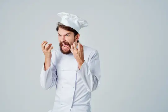 Разъяренный повар в колпаке сжимает кулаки на сером фоне