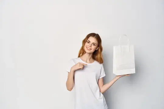 Улыбающаяся рыжеволосая девушка указывает на белый бумажный пакет