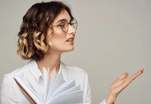 Молодая женщина в очках с книгой жестикулирует на сером фоне