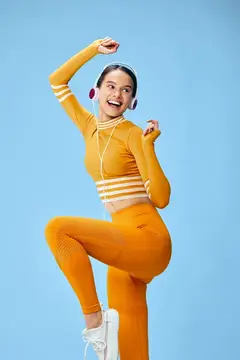 Улыбающаяся девушка в оранжевом спортивном костюме и наушниках танцует на голубом фоне