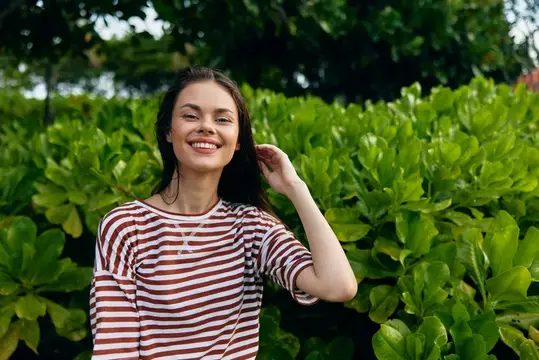 Улыбающаяся темноволосая девушка в полосатой футболке на фоне зелёных кустов
