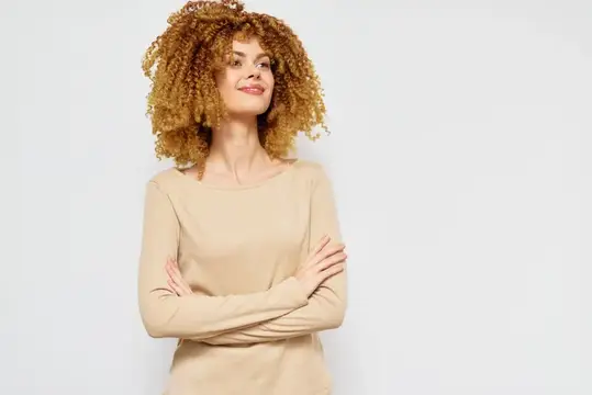 Улыбающаяся женщина с вьющимися волосами и естественным макияжем, одетая в бежевый топ, со скрещёнными на груди руками на нейтральном цветном фоне, олицетворяет красоту и уверенность в себе в…