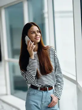 Счастливая женщина улыбается, стоя у окна, и разговаривает по телефону
