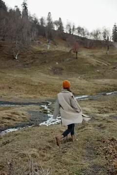 Женщина в оранжевой шапке идёт по осеннему лугу к ручью
