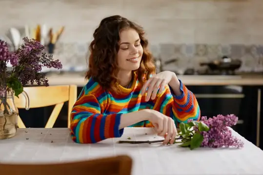 Улыбающаяся кудрявая девушка в радужном свитере за кухонным столом с сиренью