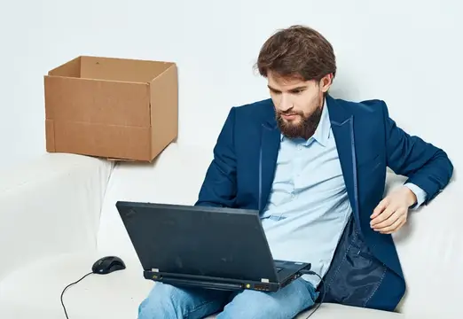 Бородатый мужчина в синем пиджаке работает за ноутбуком на диване