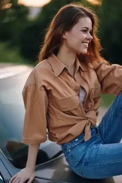 Улыбающаяся девушка в рубашке сидит на капоте автомобиля