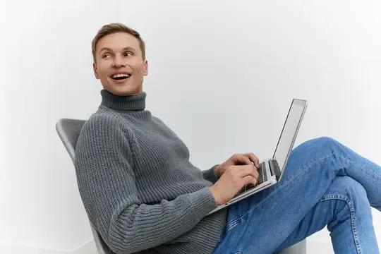Улыбающийся мужчина в сером свитере работает за ноутбуком в кресле