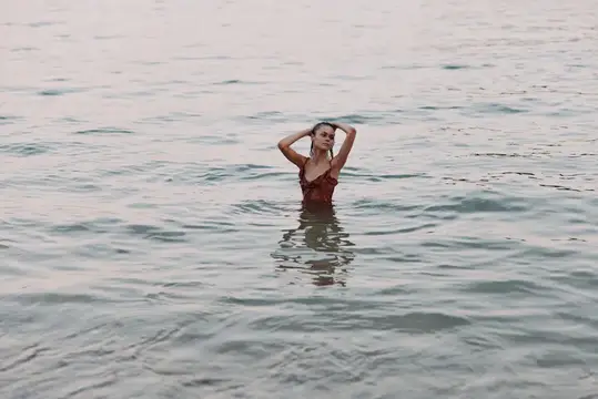 Молодая женщина в красном платье спокойно стоит в нежной воде на закате, выражая умиротворение и связь с природой в мирной обстановке у моря