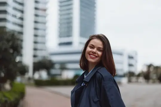 Молодая женщина с темными волосами улыбается в камеру на открытом воздухе в городской обстановке