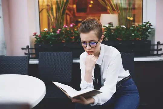 Молодой человек в очках задумчиво читает книгу за столиком кафе