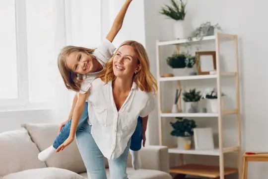 Светловолосая мама несёт на спине смеющуюся дочку в светлой гостиной