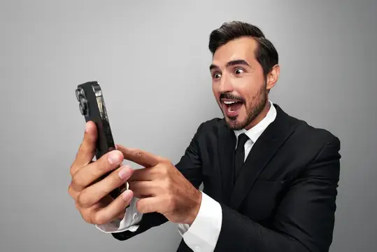 Удивлённый мужчина в костюме смотрит в смартфон на сером фоне