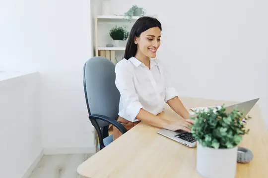 Улыбающаяся темноволосая женщина работает за ноутбуком в светлом офисе