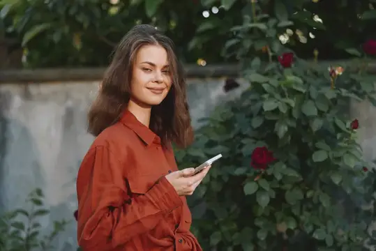 Молодая женщина с телефоном улыбается на фоне розового куста