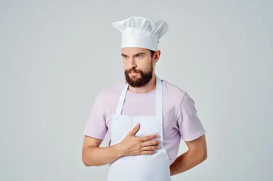 Бородатый мужчина в поварском колпаке и фартуке прижимает руку к груди