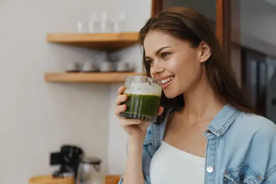 Улыбающаяся темноволосая женщина пьёт зелёный смузи на кухне