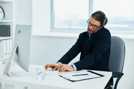 Мужчина в очках и костюме говорит по телефону за компьютером в офисе