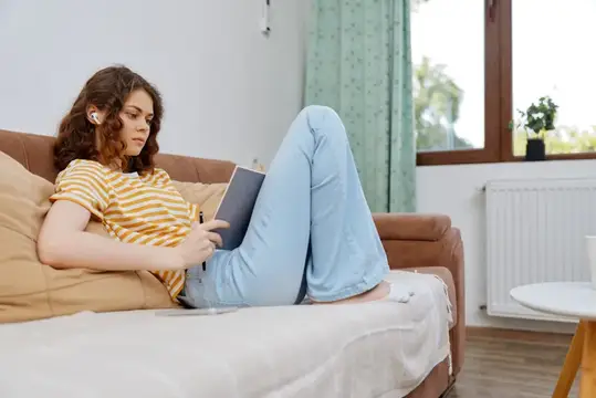 Кудрявая девушка в наушниках читает книгу на диване