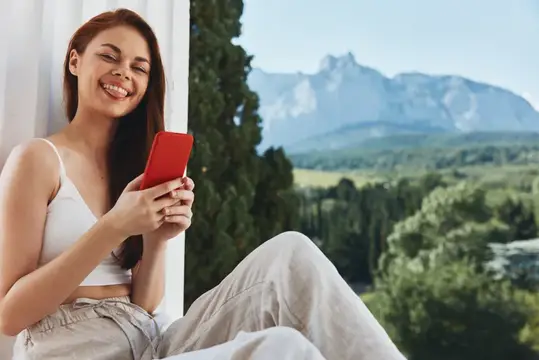 Улыбающаяся девушка с красным смартфоном на фоне гор