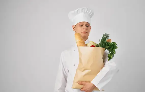Повар в белом колпаке держит бумажный пакет с багетом и зеленью