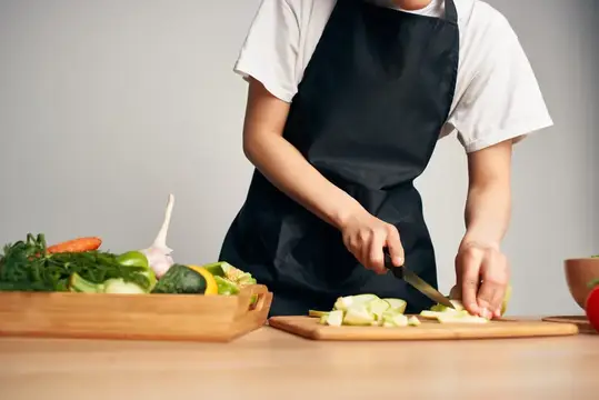 Повар в чёрном фартуке нарезает овощи на кухне