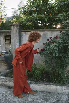 Кудрявая женщина в терракотовом костюме нюхает розы у стены