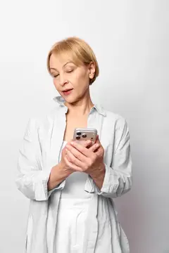 Светловолосая женщина в рубашке смотрит в смартфон на светлом фоне
