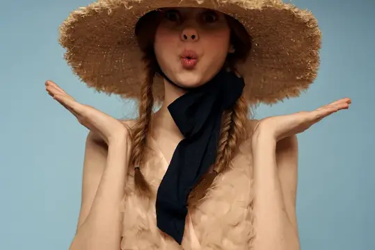 Молодая женщина в соломенной шляпе с косичками дует губы на голубом фоне