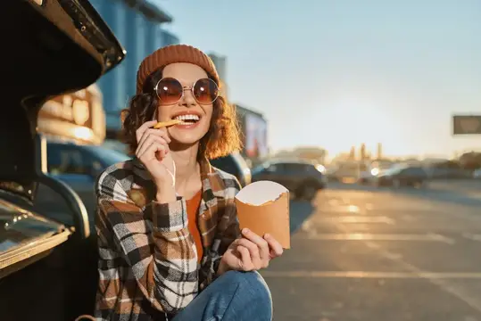 Улыбающаяся девушка в круглых очках ест картошку фри у машины