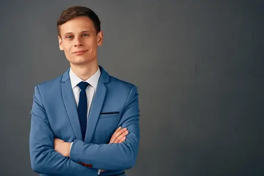 Молодой человек в синем костюме со скрещёнными руками на сером фоне